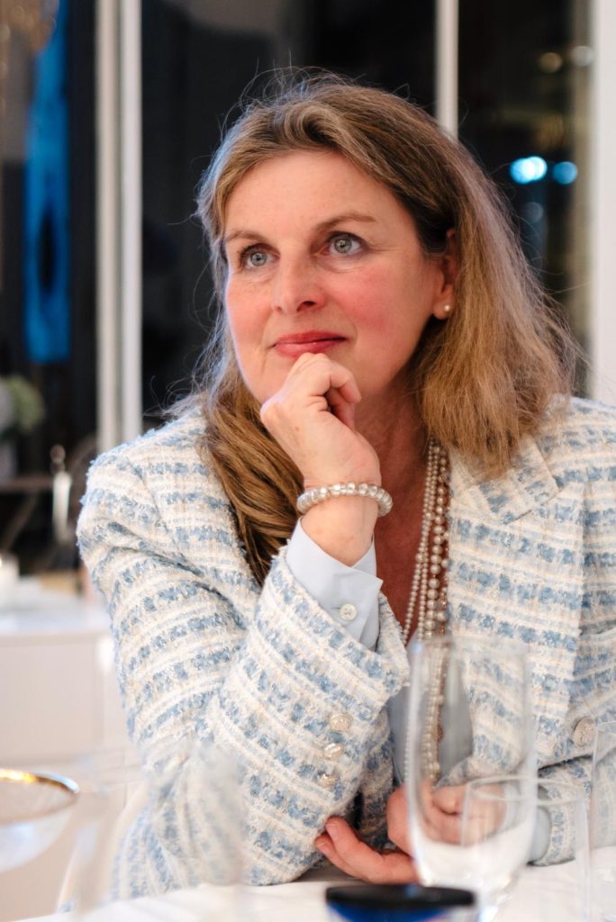 Eine Frau in hellblau-weißer Jacke sitzt am Tisch, stützt das Kinn auf eine Hand und blickt zur Seite.
