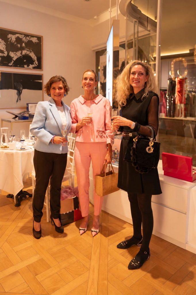 Drei Frauen in hellblauer, rosa und schwarzer Kleidung stehen mit Gläsern zusammen im Salon.