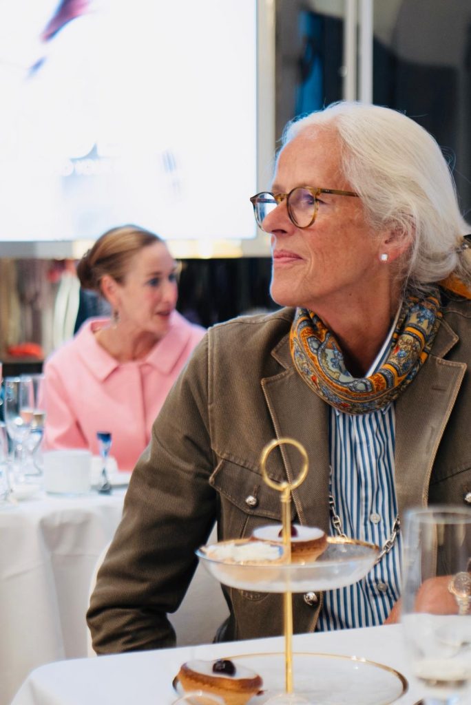 Eine Frau mit weißen Haaren, Brille und buntem Schal blickt am gedeckten Tisch zur Seite; vor ihr steht eine Etagere.