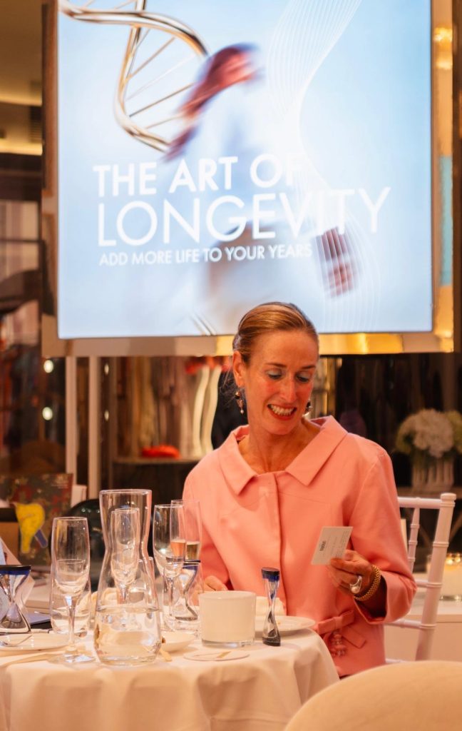 Eine Frau in Rosa sitzt an der gedeckten Tafel; hinter ihr zeigt ein Bildschirm „The Art of Longevity“.