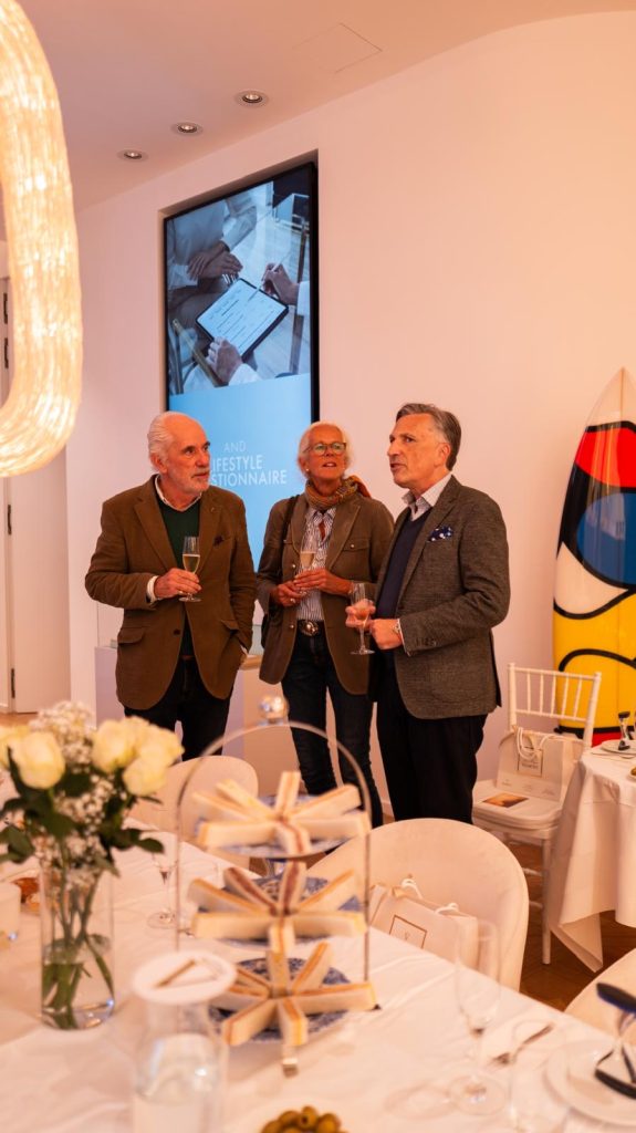 Drei Männer stehen mit Gläsern hinter einer gedeckten Tafel mit Blumen und Etageren.