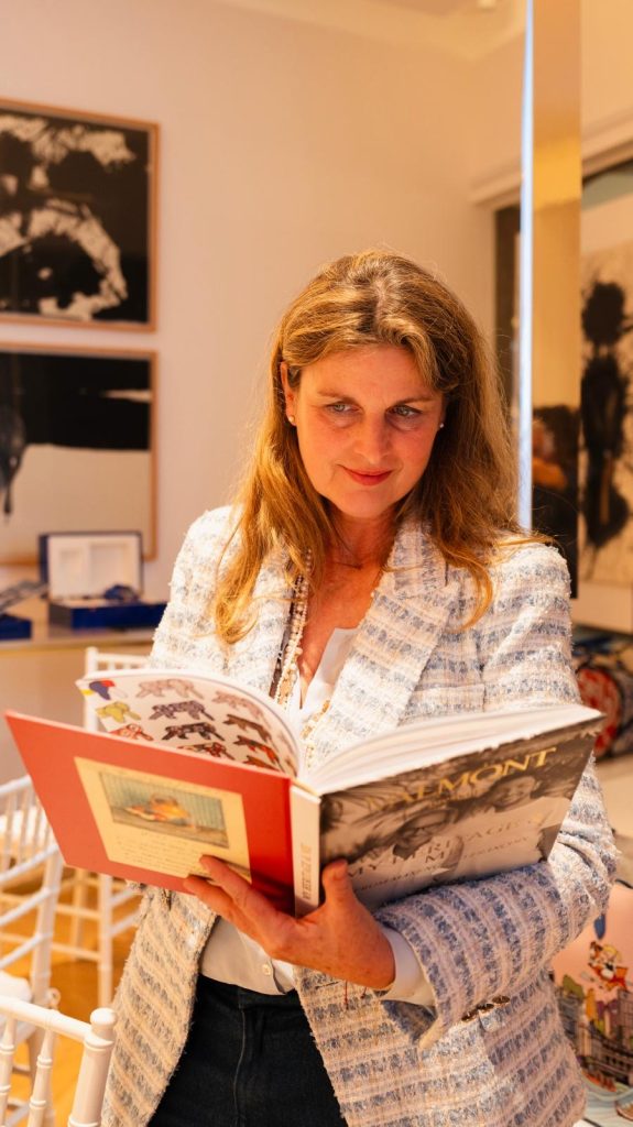 Eine Frau in einer hellblau-weißen Jacke hält ein aufgeschlagenes Valmont-Buch und blickt zur Seite.