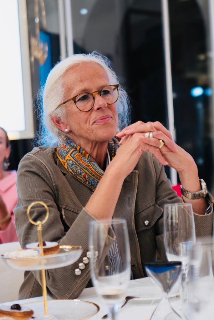 Eine Frau mit weißen Haaren, Brille und buntem Schal sitzt mit gefalteten Händen an der Tafel.