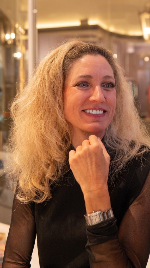 Eine Frau mit lockigem blondem Haar lächelt am Tisch und hält eine Hand unter ihr Kinn.